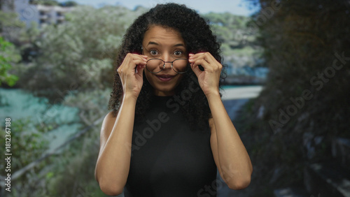 Obraz na plátně Smiling woman with curly hair wearing glasses in a lush park setting engages naturally outdoors showing joy and confidence