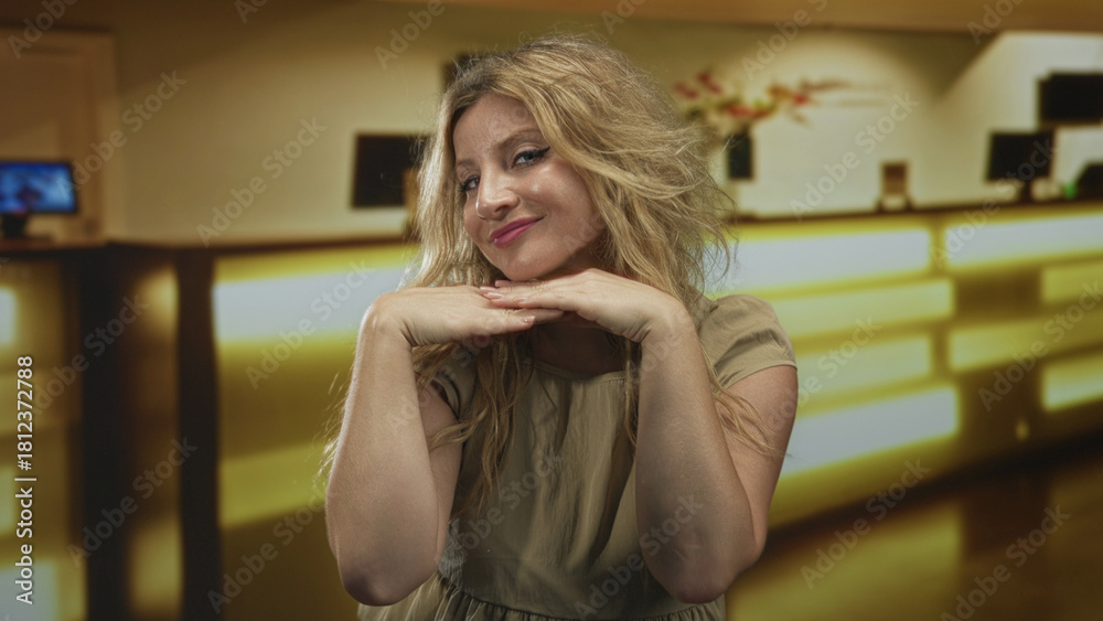 Fototapeta premium Blonde woman resting chin on hands in hotel lobby smiling at camera with soft golden lighting and reception desk visible; confidence.
