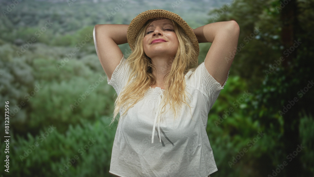 Naklejka premium Woman wearing straw hat and white blouse with arms raised in forest among green foliage, smiling with eyes closed; serenity escape.