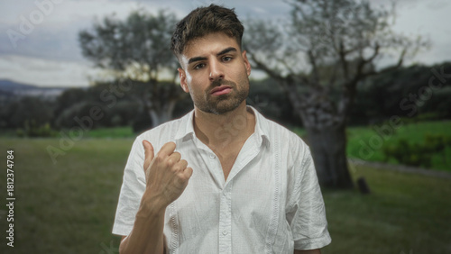 Bearded young man in white shirt points thumb to sunlit green forest clearing while gazing sideways; uncertainty.