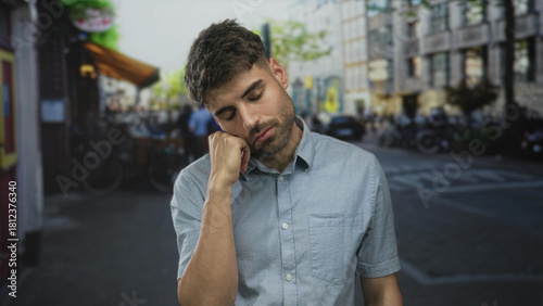 Fotografie Man with eyes closed leaning head on hand on quiet urban street corner lined with old buildings; weariness solitude introspection boredom