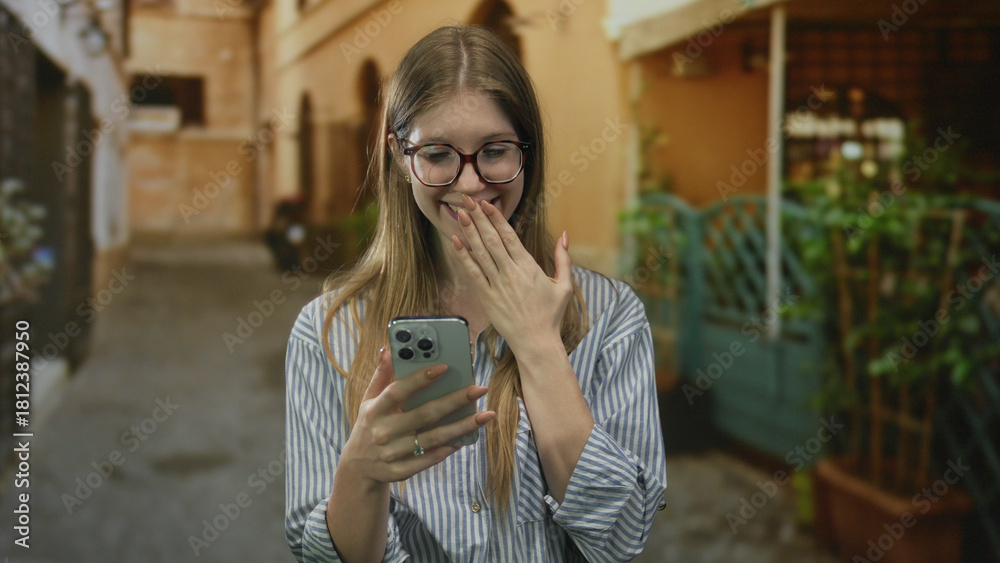 Obraz premium Woman covers mouth with hand while looking at smartphone screen on a sunlit old town street; surprise.