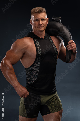 strong athlete stands proudly with a weight bag resting on his shoulder. He showcases defined muscles while dressed in athletic gear. The background is dark highlighting his physique.