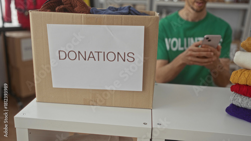 Man holds smartphone to ear at donation center table with a large cardboard box labeled donations and a stack of colorful sweaters; compassion.