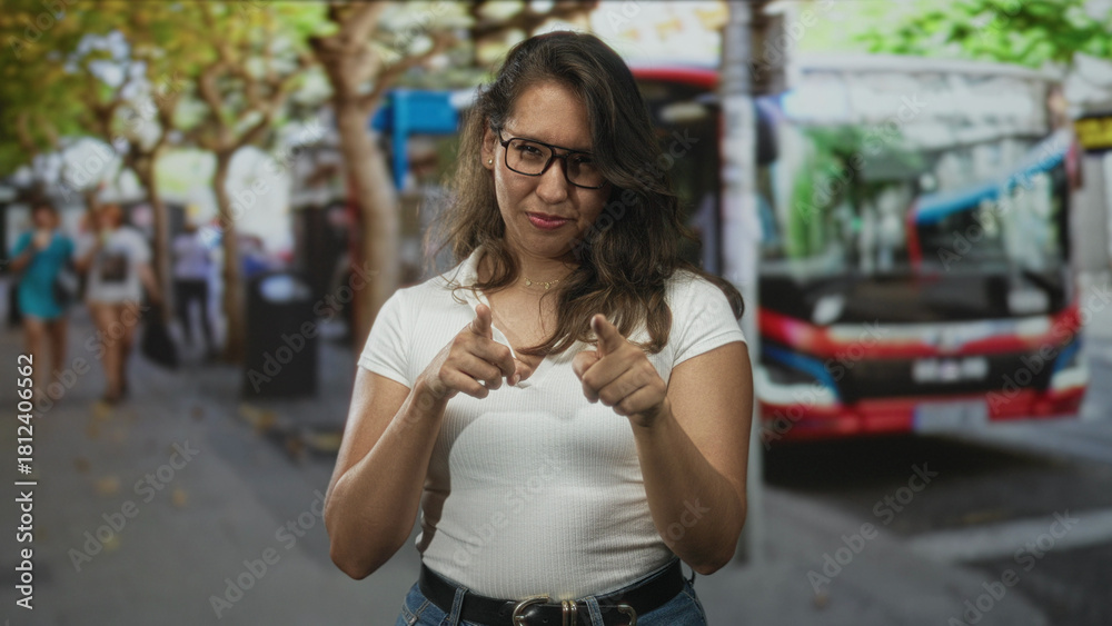 Fototapeta premium Woman points finger to camera on busy city street while smiling and wearing glasses and white top; playful friendly confidence.
