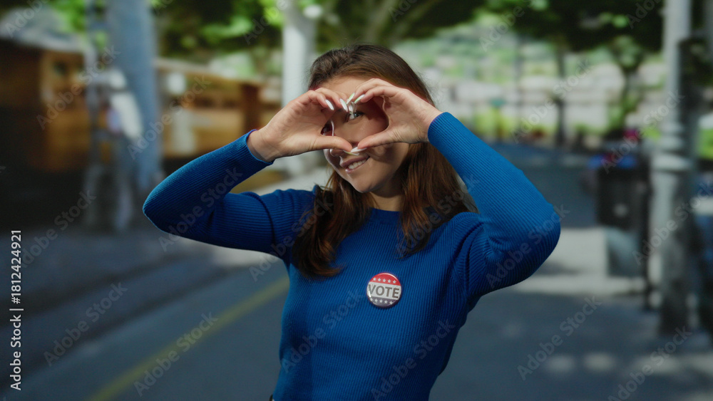 Fototapeta premium Woman wearing vote badge forms heart shape with hands on city street smiling in sunlight during us election outdoors showing patriotic support