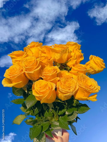 A vibrant bouquet of fresh yellow roses held up against a bright blue sky with soft white clouds. 