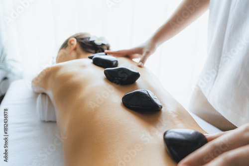 Hot stone massage at spa salon in luxury resort with day light serenity ambient, blissful woman customer enjoying spa basalt stone massage glide over body with soothing warmth. Quiescent