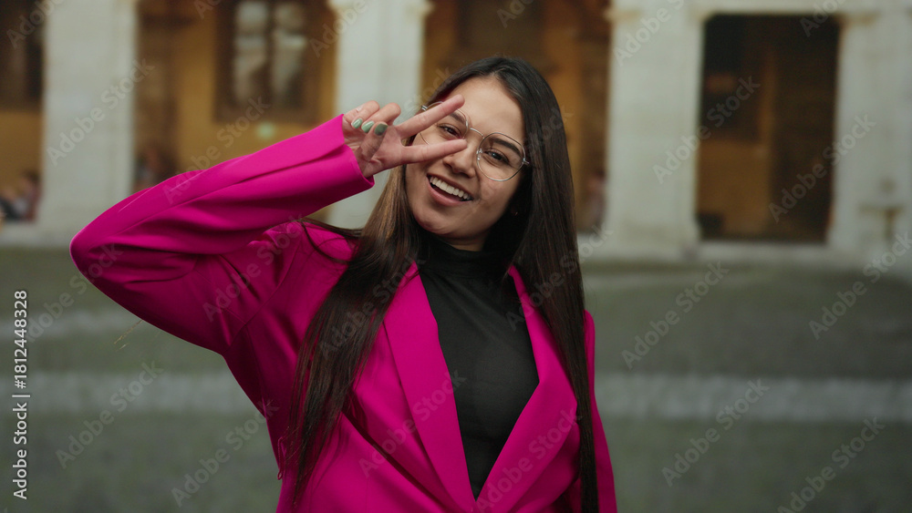 Fototapeta premium Woman smiling outside historic university wearing pink suit and glasses with a peace sign gesture, capturing youthful energy in an educational setting.