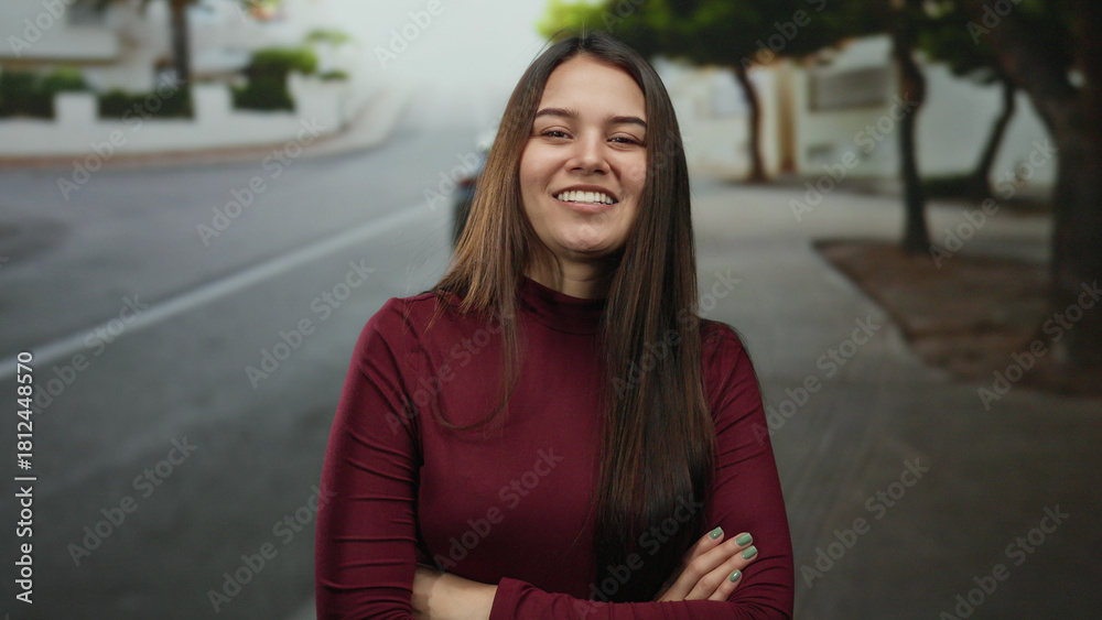 Fototapeta premium Woman smiling with arms crossed standing on an outdoor street, showcasing a confident and relaxed expression in a casual urban environment.