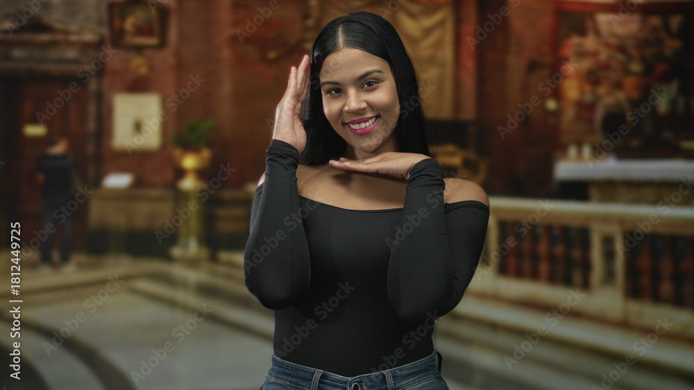 Fototapeta premium Smiling young latin woman frames her face with hands against ornate stone building interior walls; happiness.