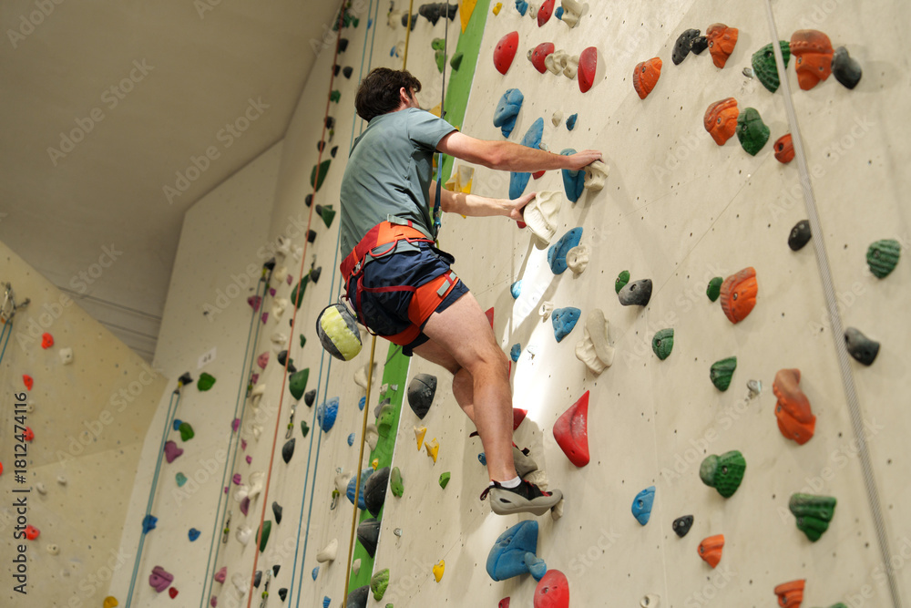 Fototapeta premium man with safety rope and alpine harness climbing indoor wall with colorful holds