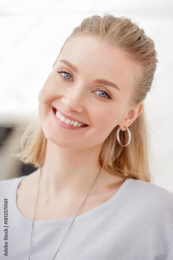 Naklejka premium A beautiful blonde woman smiles warmly, wearing a casual gray top and large hoop earrings. A joyful and inviting portrait.