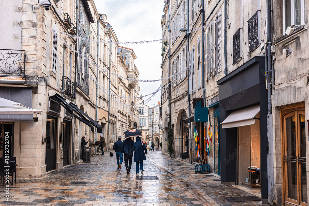 Obraz premium A charming narrow street in La Rochelle featuring historic limestone facades and traditional arcades. An artisan bakery and cafe tables line the wet cobblestone pavement under an overcast sky.