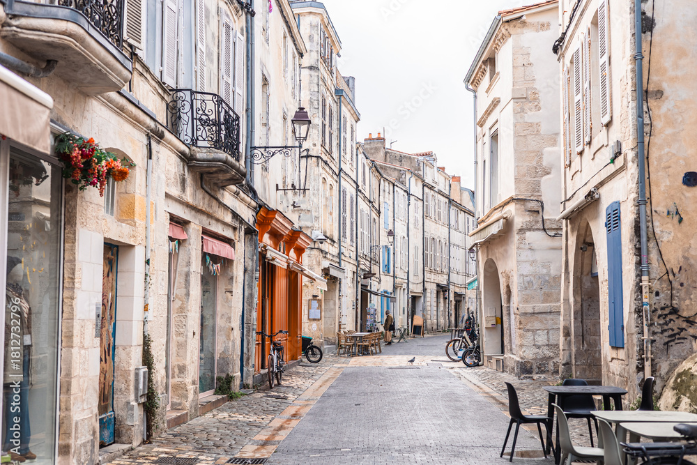 Obraz premium A charming narrow street in La Rochelle featuring historic limestone facades and traditional arcades. An artisan bakery and cafe tables line the wet cobblestone pavement under an overcast sky.