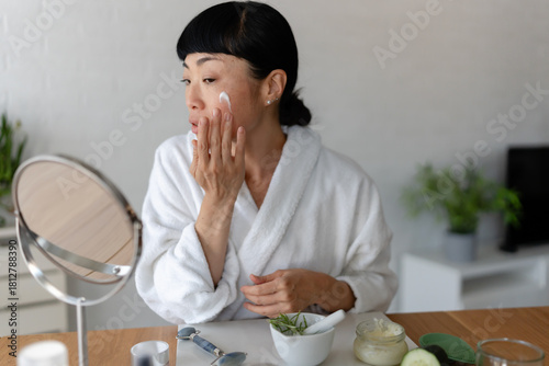 Smiling Asian woman in a white bathrobe applying face cream while looking into a mirror, surrounded by natural skincare products in a relaxed home setting