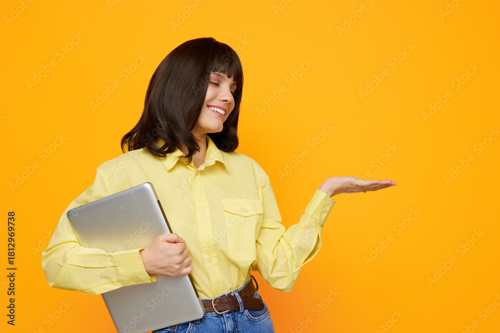 Naklejka premium A cheerful woman in a yellow shirt smiles while holding a laptop against a vibrant orange background, creating a friendly, approachable office or study scene.