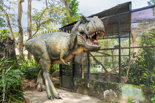 Realistic life-size Tyrannosaurus rex dinosaur replica in an outdoor park exhibit surrounded by trees and vegetation