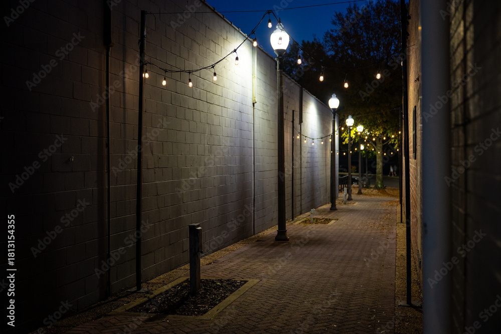 Fototapeta premium Moody nighttime alleyway illuminated by street lamps and hanging string lights, with brick paving and deep blue sky. A cinematic urban scene ideal for travel, architecture, and atmospheric design them