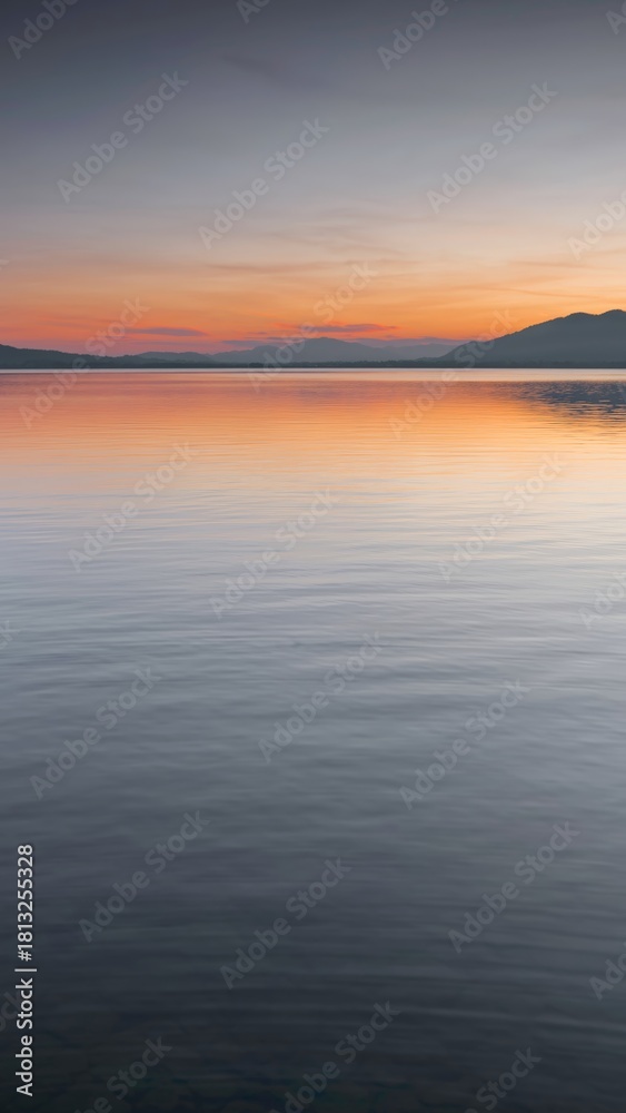 Fototapeta premium Calm lake at sunset with distant mountains and colorful sky.