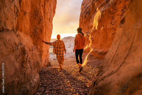 Two young women tourists are standing at lake or sea beach between big rocks at the exit from narrow canyon and looks at sunset shore