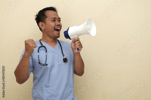 Southeast Asian man in medical scrubs with stethoscope shouting into megaphone, promoting healthcare message or campaign; isolated with copy space for advertisement.
