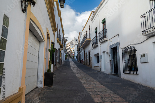 Fototapeta Naklejka Na Ścianę i Meble -  A narrow, cobblestone street in Nijar, Almeria, Spain, lined with whitewashed buildings, wooden doors, small balconies, and potted plants