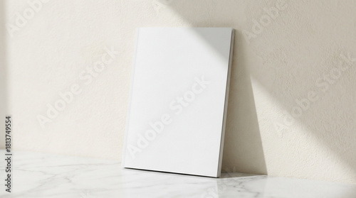 Blank White Book Mockup with a Clean Cover Leaning Against a Textured Beige Wall, Resting on a Polished Marble Surface, Bathed in Sunlight.