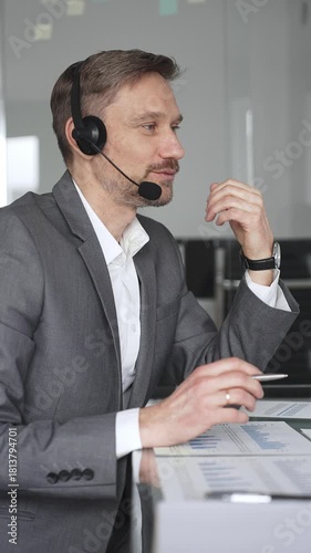 Businessman professional wearing a headset, engaging in an online video call or teleconference, discussing financial data and charts while working in a modern office setup, vertical view