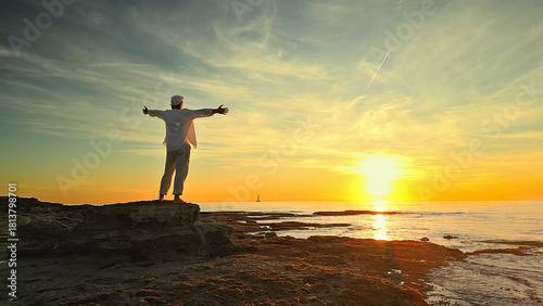A person stands on a rocky shore at sunset, arms outstretched, embodying mindfulness and reflection. The serene ocean and golden sky evoke peace and escape, promoting mental wellbeing.
