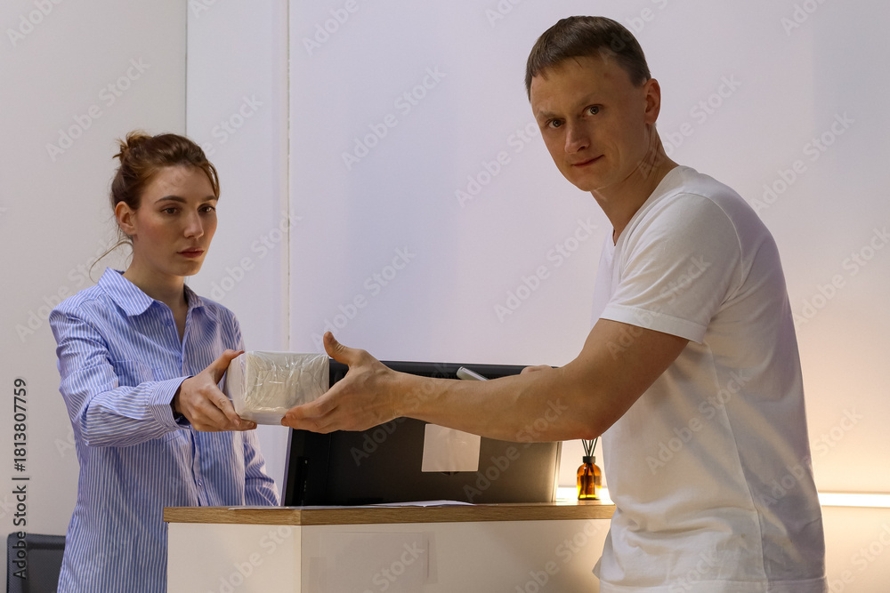 Obraz premium Woman handing small wrapped package to man at counter. Female clerk in blue striped shirt passes compact parcel to male customer in white shirt across wooden counter with monitor