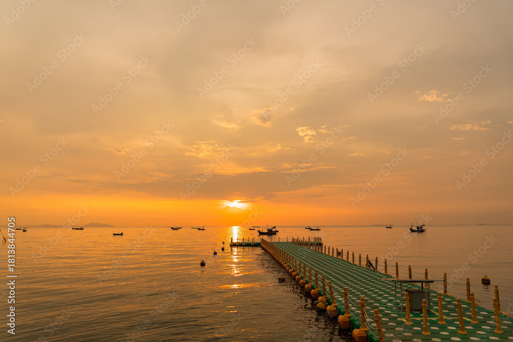 Obraz premium Floating dock extending into calm sea during golden sunrise with fishing boats on the horizon under a colorful sky