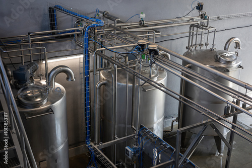The interior of a modern industrial plant, such as a food, beverage, or chemical factory. Large stainless steel tanks are connected by a complex system of pipes, valves, and sensors.