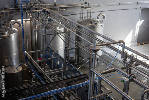 The interior of a modern industrial plant, such as a food, beverage, or chemical factory. Large stainless steel tanks are connected by a complex system of pipes, valves, and sensors.