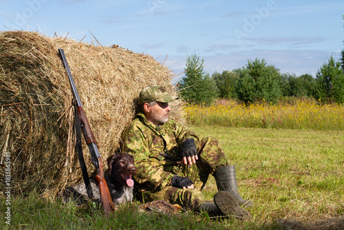 A hunter and his dog relax in the shade of a hay bale