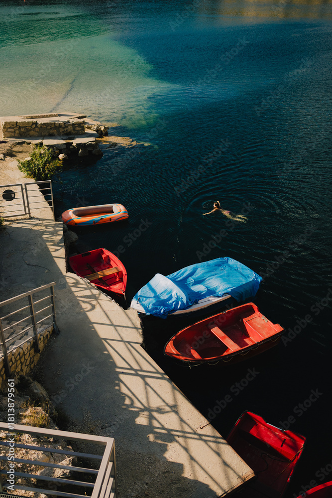 Fototapeta premium Woman swimming in stunning lake, surrounded by colorful boats moored at pier, savoring warmth of sunny summer day while enjoying tranquility of nature. Female gliding, clear lake, colorful boats