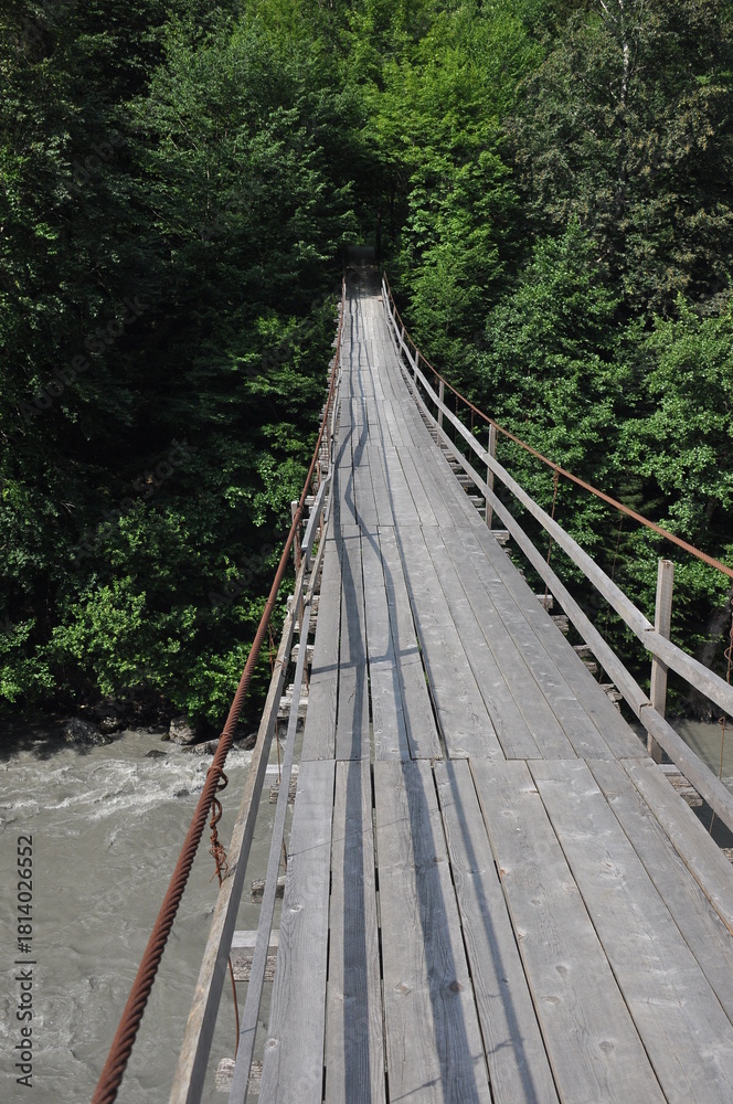 Obraz premium Wooden suspension bridge stretches over a river surrounded by lush greenery in summer