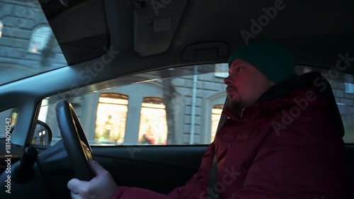 Man driving a car, wearing a beanie and red jacket. Street view visible through the window
