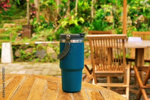 A blue travel tumbler standing on a wooden outdoor table with chairs and garden scenery, ideal for lifestyle and product concepts.