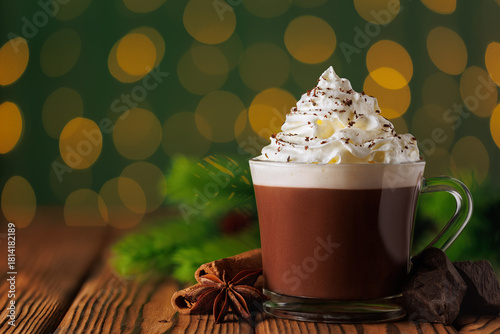 Glass mug of hot chocolate with whipped cream and cinnamon sticks against green background of bokeh glowing garland lights