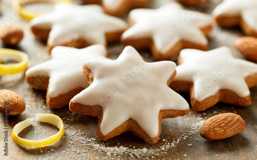 A close-up photograph of traditional German Zimtsterne (cinnamon stars) arranged on a rustic wooden surface, showcasing their distinctive star shape and glossy white icing glaze. The delicate cookies 