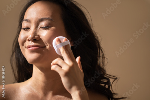 Asian woman applying skincare product with a cusion cover, showcasing a radiant complexion and natural beauty, in a softly lit environment emphasizing self-care and wellness