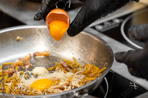 A chef adds a fresh egg yolk to a creamy carbonara pasta dish in a stainless steel pan, highlighting the cooking process.