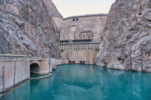 Massive concrete Toktogul HPP dam in a narrow rocky canyon. Turquoise water below, tunnel on the left. High stone walls, clear sky.