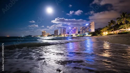 A moonlit beach at night. Buildings with lights line the shore, palm trees frame the right, and waves crash against the sand