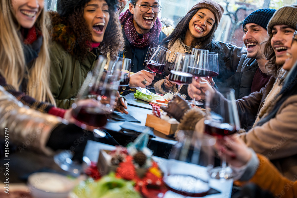 Naklejka premium Happy multiracial friends toasting red wine at restaurant terrace - Group of young people wearing winter clothes having fun at outdoors winebar table - Dining life style and friendship concept
