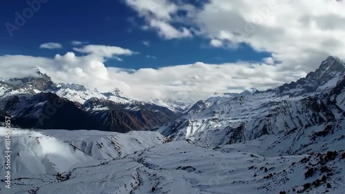 A panoramic vista unveils a snow-draped mountain range beneath a vibrant blue sky dotted with fluffy clouds. Snowy valleys lie below peaks