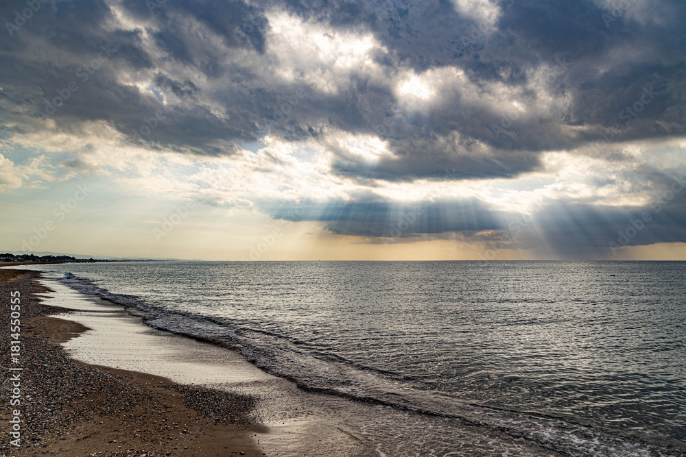 Obraz premium A deserted Mediterranean beach under a cloudy sky, where sunbeams break through the clouds to illuminate the calm sea. A peaceful interseason moment on the shore in Sorgun, Antalya, Turkey.
