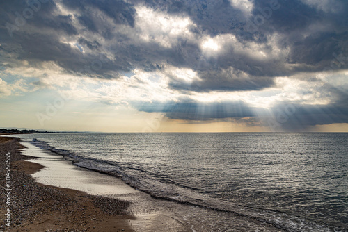 Fototapeta Naklejka Na Ścianę i Meble -  A deserted Mediterranean beach under a cloudy sky, where sunbeams break through the clouds to illuminate the calm sea. A peaceful interseason moment on the shore in Sorgun, Antalya, Turkey.

