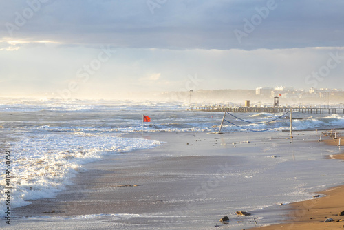 Fototapeta Naklejka Na Ścianę i Meble -  Stormy sea with massive waves and white foam under bright sunlight. Tilted volleyball poles stand in surf as red storm flag waves in the turbulent water. Side, Antalya, Turkey, Mediterranean.

